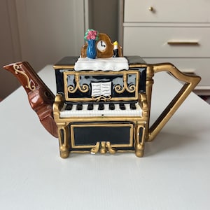 May include: A decorative teapot shaped like a piano, featuring a black and gold design with a keyboard and music sheet details. The teapot has a brown spout and a gold harp-shaped handle. A small clock, candle, and vase of flowers adorn the top.