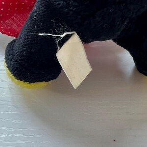 May include: Close-up of a black plush toy with a red polka dot bow and yellow accents. A blank, rectangular tag is attached to the toy. The toy's fur is black and textured.
