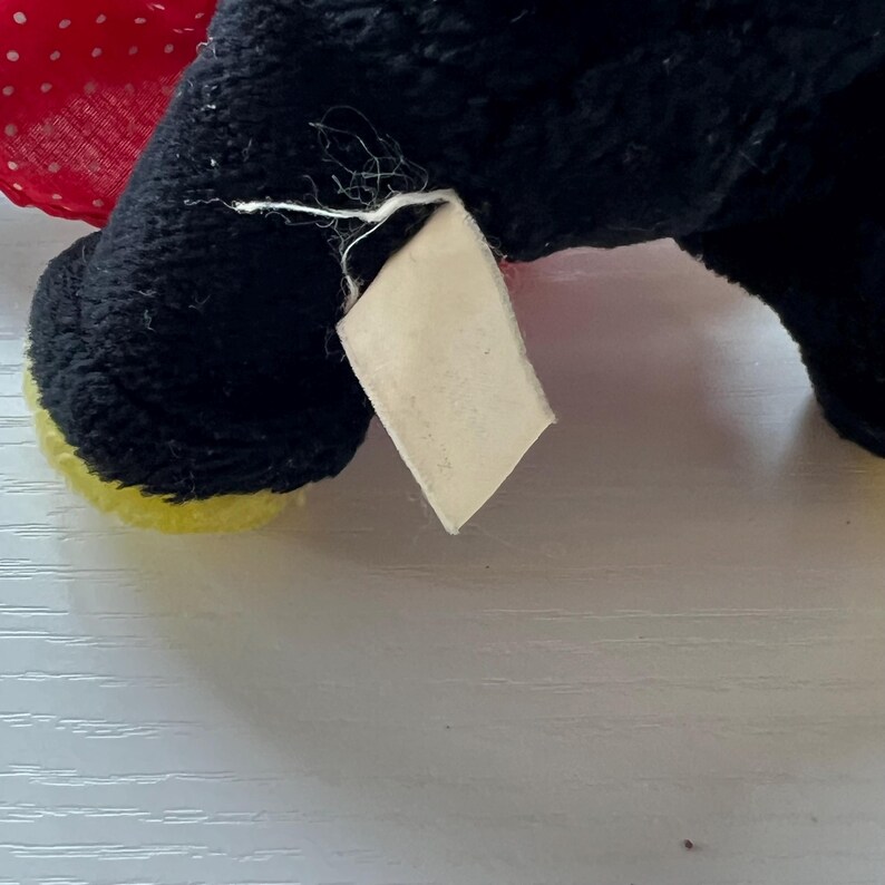 May include: Close-up of a black plush toy with a red polka dot bow and yellow accents. A blank, rectangular tag is attached to the toy. The toy's fur is black and textured.