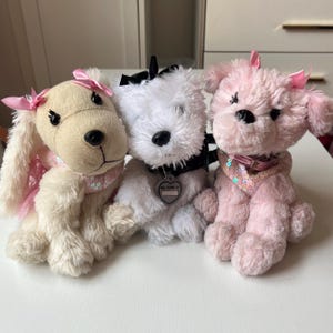 May include: Three plush dog toys in cream, white, and pink. Each dog has a bow, collar, and a sequined scarf. The white dog has a tag that says "My name is". The toys are soft and fluffy.