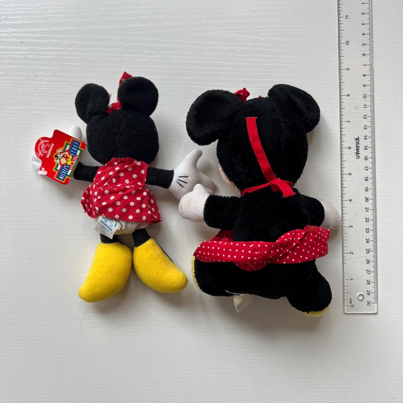 May include: Two Minnie Mouse plush toys. One is facing forward, holding a tag, and the other is facing away. Both have black fur, red and white polka dot dresses, white gloves, yellow shoes, and red bows.