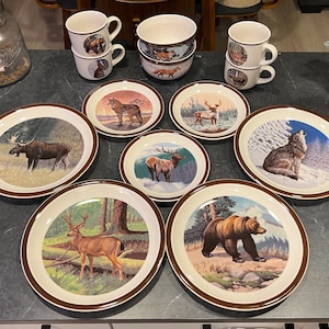 以下が含まれることがあります： 野生動物のシーンが特徴の、ヴィンテージセラミック食器のコレクション。セットには、クマ、シカ、オオカミなどの動物の画像でそれぞれ装飾された、プレート、ボウル、マグカップが含まれています。プレートには茶色の縁があり、マグカップにはハンドルが付いています。