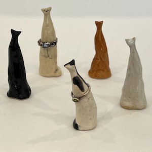 May include: Five ceramic cat-shaped ring holders in various colors including black, cream, brown, and a speckled white with black patches. Two of the figures are wearing silver rings around their necks, one with a small green stone.