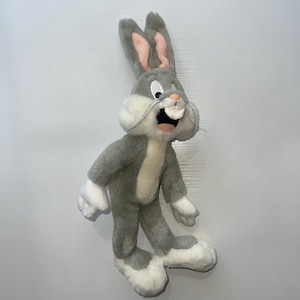 Puede incluir: Un peluche de Bugs Bunny en posición de pie. El icónico personaje de dibujos animados es principalmente gris con detalles blancos en el pecho, las patas y los pies. El interior de las orejas es rosa y la boca está abierta, mostrando un gran diente.