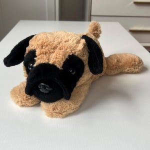 May include: A plush pug dog toy with tan fur and black accents on the ears, snout, and around the eyes. The toy is lying down on a white surface, with its paws extended. The pug has a soft, cuddly appearance.