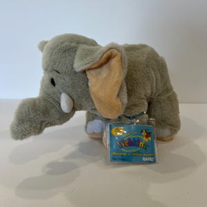 May include: A gray plush elephant toy with large tan inner ears and white tusks. A clear plastic tag is attached to the side, featuring the text Webkinz, discover a virtual world, and GANZ, along with a small illustration of a dog.