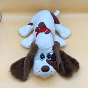 Pode incluir: Um brinquedo de pelúcia de cachorro com corpo branco, manchas marrons e orelhas marrons longas. O cão tem olhos azuis grandes, um nariz marrom e uma coleira vermelha. O brinquedo está deitado em uma superfície amarelo claro.