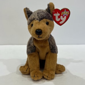 May include: A plush dog toy with tan and dark gray fur, featuring black plastic eyes and a black nose. A red heart-shaped tag is attached to the ear, displaying the white text ty and the year 2000 with a silver star graphic on the front.