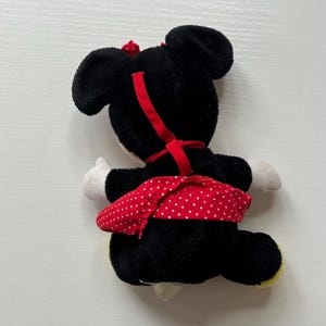 May include: A Minnie Mouse soft toy, seen from behind, with black fur, white gloves, and a red polka-dot skirt. A red bow sits on the head, and a red ribbon runs down the back. The toy has yellow feet.