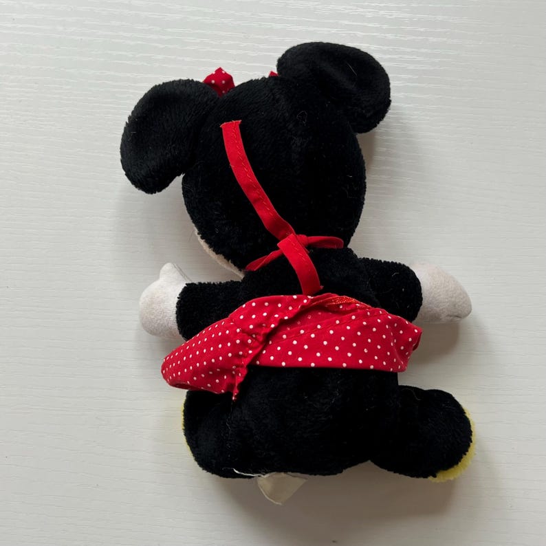 May include: A Minnie Mouse soft toy, seen from behind, with black fur, white gloves, and a red polka-dot skirt. A red bow sits on the head, and a red ribbon runs down the back. The toy has yellow feet.