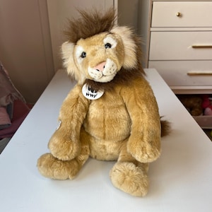 Puede incluir: Un león de peluche con cuerpo marrón, melena marrón y nariz rosa. El león tiene una etiqueta blanca con el logo de WWF. El juguete está sobre una superficie blanca. El pelaje del león es suave y esponjoso.