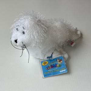 May include: A white plush seal toy with a shaggy mane and black whiskers. The toy has a Webkinz tag attached, featuring a blue background and the words "Webkinz Discover a virtual world!"