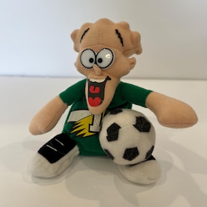 May include: A plush toy character with tan skin, large white eyes, and a wide open mouth, wearing a green shirt with a yellow lightning bolt and the number one. The character is sitting down and holding a white soccer ball with black pentagon patches.