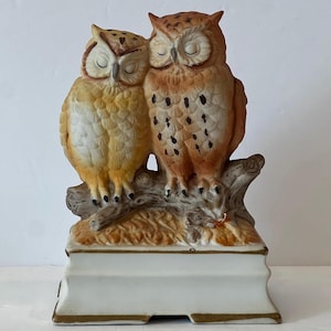 May include: A ceramic figurine featuring two owls with closed eyes perched together on a textured tree branch. The owls have yellow and brown feathers with dark brown spots. The base is white with gold trim along the bottom and top edges.
