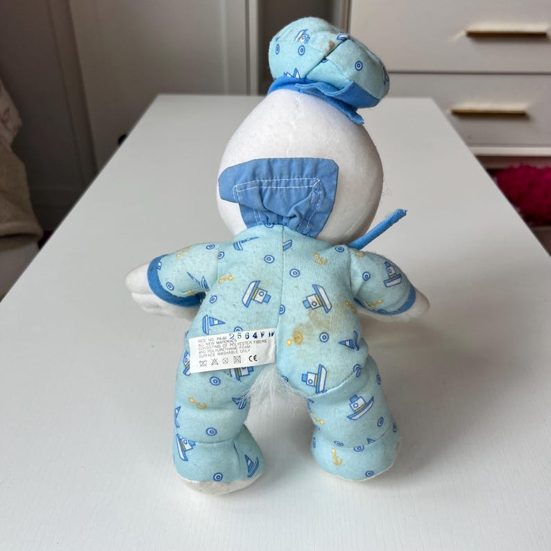 May include: A plush toy in the shape of a sailor, viewed from the back. The toy is mostly white with blue accents. It wears a blue sailor hat and a light blue onesie patterned with boats and anchors. The toy is on a white surface.