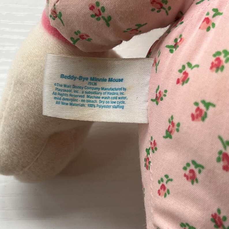 May include: Close-up of a Beddy-Bye Minnie Mouse plush toy. The toy has a pink floral patterned fabric and a white tag with text. The tag reads "Beddy-Bye Minnie Mouse" and includes washing instructions.