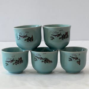 May include: A set of five light blue ceramic tea cups, each featuring a dark brown graphic illustration of a bird in flight above a pine tree and small clouds. The cups have a smooth, rounded shape with a wide rim and a solid base.