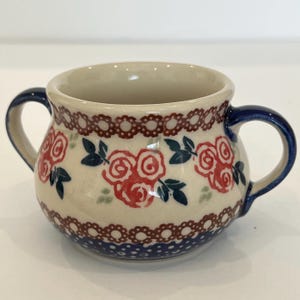 May include: A cream-colored ceramic sugar bowl with two handles, featuring a hand-painted pattern of red roses with dark blue leaves, accented by a brown scalloped border and a dark blue base with small white dots around the bottom edge.