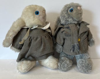 May include: Two plush rabbit toys with blue eyes. The left rabbit is cream-colored, wearing a brown and white houndstooth dress with a white collar. The right rabbit is gray, wearing a matching brown and white houndstooth jacket and a fringed scarf.