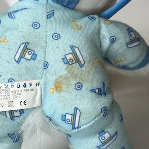 May include: Close-up of a plush toy's back, featuring light blue fabric patterned with boats, anchors, and circles. The toy has a white fluffy area and a tag with text. Some brown stains are visible on the fabric.