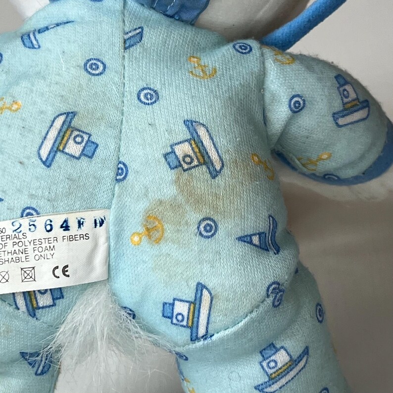 May include: Close-up of a plush toy's back, featuring light blue fabric patterned with boats, anchors, and circles. The toy has a white fluffy area and a tag with text. Some brown stains are visible on the fabric.