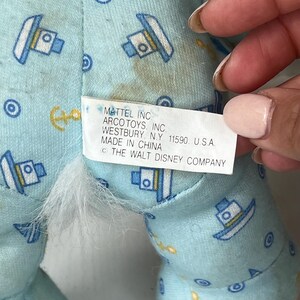 May include: Close-up of a light blue fabric with a pattern of boats, anchors, and circles. A white tag with black text reads "MATTEL INC ARCO TOYS, INC. WESTBURY, N.Y. 11590. U.S.A. MADE IN CHINA (C) THE WALT DISNEY COMPANY."