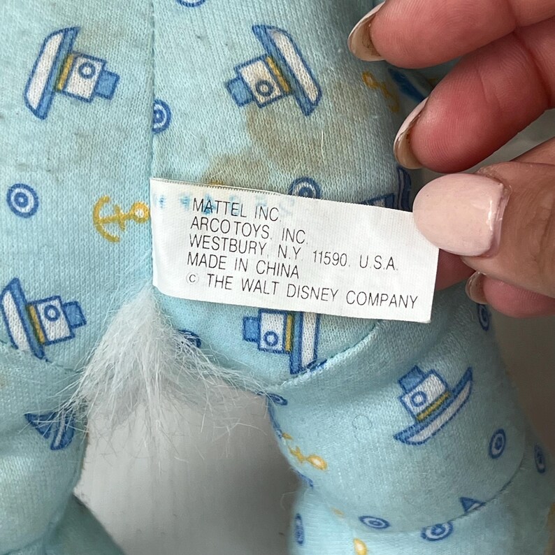 May include: Close-up of a light blue fabric with a pattern of boats, anchors, and circles. A white tag with black text reads "MATTEL INC ARCO TOYS, INC. WESTBURY, N.Y. 11590. U.S.A. MADE IN CHINA (C) THE WALT DISNEY COMPANY."