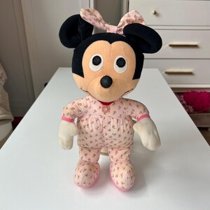 May include: A plush Minnie Mouse doll wearing a pink floral print pajama set and bow. The doll has black ears, a black nose, and white gloves. The doll is standing on a white surface.