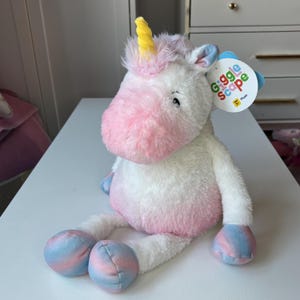 Pode incluir: Um brinquedo de unicórnio de pelúcia com corpo branco, focinho rosa e cascos azuis e rosa. O unicórnio tem um chifre amarelo e uma etiqueta que diz "Giggle Scape Plush". O brinquedo está em uma superfície branca.
