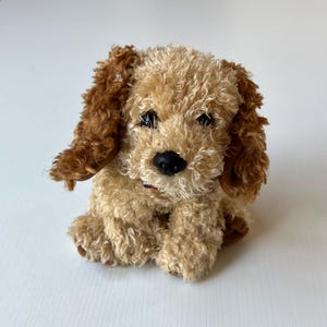 May include: A plush toy dog with tan and brown fur. The dog has floppy brown ears, a black nose, and a red collar. The toy is sitting and has a soft, textured appearance.