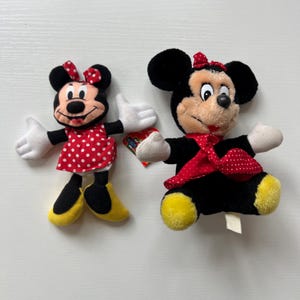 May include: Two Minnie Mouse plush toys. One wears a red polka dot dress and bow, the other a red polka dot scarf and bow. Both have black ears, black bodies, white gloves, and yellow shoes. The toys are on a white surface.