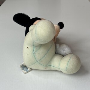 May include: A plush Mickey Mouse toy, featuring black ears, a black nose, and a cream-colored face. The body is a light green color with blue stitching. The toy is lying on its side.