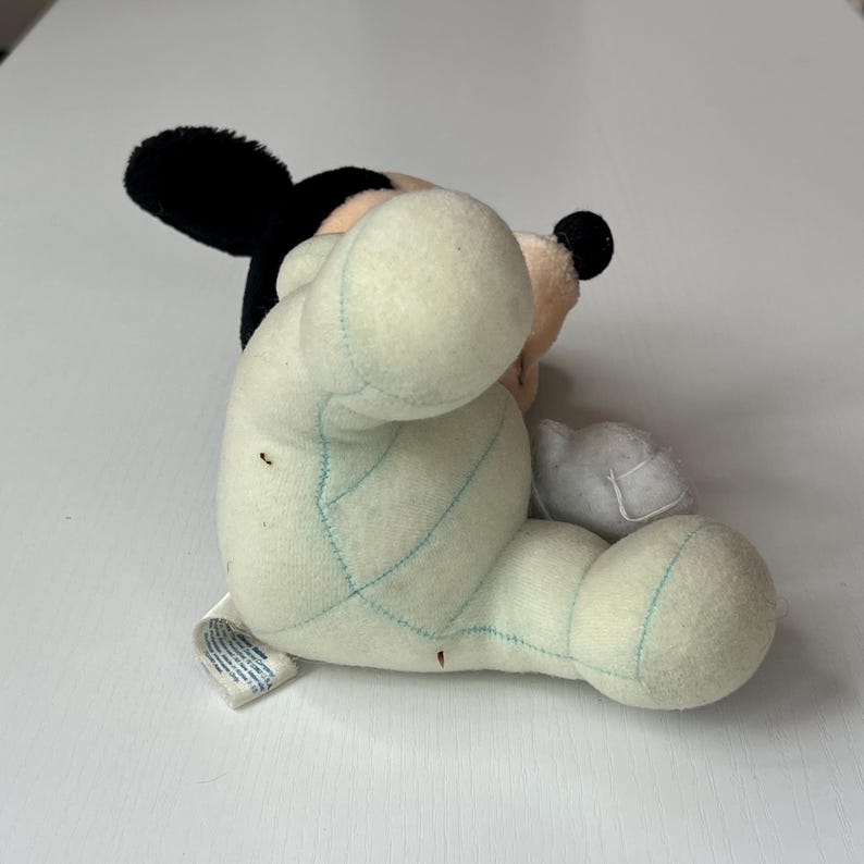 May include: A plush Mickey Mouse toy, featuring black ears, a black nose, and a cream-colored face. The body is a light green color with blue stitching. The toy is lying on its side.
