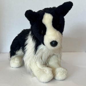May include: A plush stuffed animal designed to resemble a border collie dog. The toy features soft black and white fur, pointed ears, a white blaze on the face, and a black plastic nose. It is depicted in a seated position with front paws.