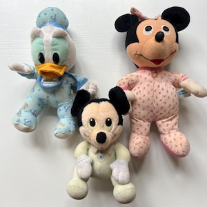 May include: Three plush Disney character dolls: Donald Duck in blue pajamas, Minnie Mouse in pink floral pajamas, and Mickey Mouse in a light green outfit. The dolls are soft and cuddly, perfect for children.