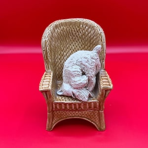 以下が含まれることがあります： 小さな茶色の籐の椅子と、白くてふわふわした犬の置物。 犬は椅子の中で丸まっており、頭と尾が見えます。 椅子は明るい赤色の背景に置かれています。