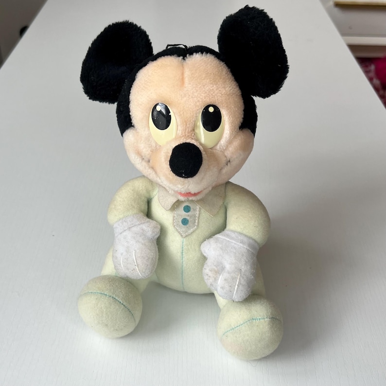 May include: A plush Mickey Mouse doll in a seated position. The doll has black ears, a black nose, and large yellow eyes. It wears a light green onesie with white gloves and light green feet. The doll has a vintage appearance.