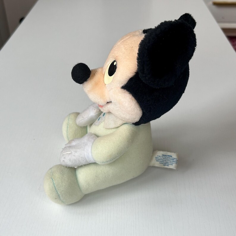 May include: A vintage Mickey Mouse plush toy in a seated position. The plush features a black head and ears, a tan face, and a light green body. The toy has white gloves and is wearing a light green outfit. A tag is attached to the bottom.