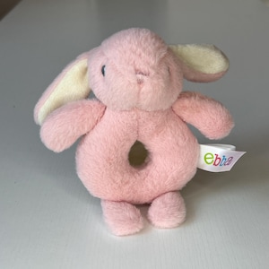 May include: A plush pink bunny rattle toy with a circular center. The bunny has long ears with cream-colored insides, small black eyes, and a tag that reads "ebba". The toy is designed for babies and young children.
