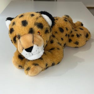 May include: A plush toy cheetah with a tan body, black spots, and a white and black face. The cheetah is lying down, with its paws extended. The toy has orange eyes and a black nose.