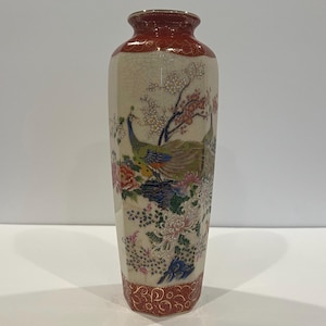 May include: A tall, hexagonal ceramic vase with a cream-colored crackle glaze body. The neck and base feature a deep red band with gold scrollwork. The body displays a colorful scene of a peacock, blooming flowers, and cherry blossom branches.