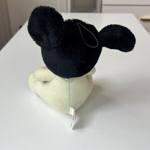 May include: A plush toy in the shape of a cartoon mouse, featuring a black head and ears, and a cream-colored body. The toy has a loop attached to the head. The toy is sitting on a white surface.