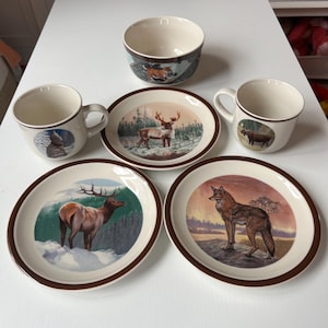 Op de afbeelding: Een verzameling crèmekleurig keramisch servies met illustraties van wilde dieren. Inclusief twee mokken, twee borden, een kom en een klein bord. De borden en de kom hebben een bruine rand. De illustraties tonen een hert, eland, wolf en andere dieren.