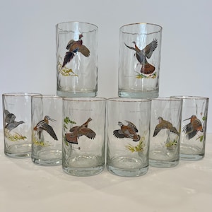 May include: A set of eight clear glass tumblers with gold-toned rims, each featuring a unique, detailed illustration of a different game bird in flight or perched among foliage, including pheasants, grouse, and woodcock, arranged in two rows.
