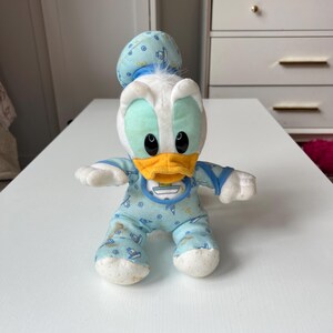 May include: A plush Donald Duck doll in a baby outfit. The doll has a white head with a blue cap, a yellow beak, and black eyes. It wears a light blue onesie with a boat pattern and a bib. The doll is approximately 8 inches tall.