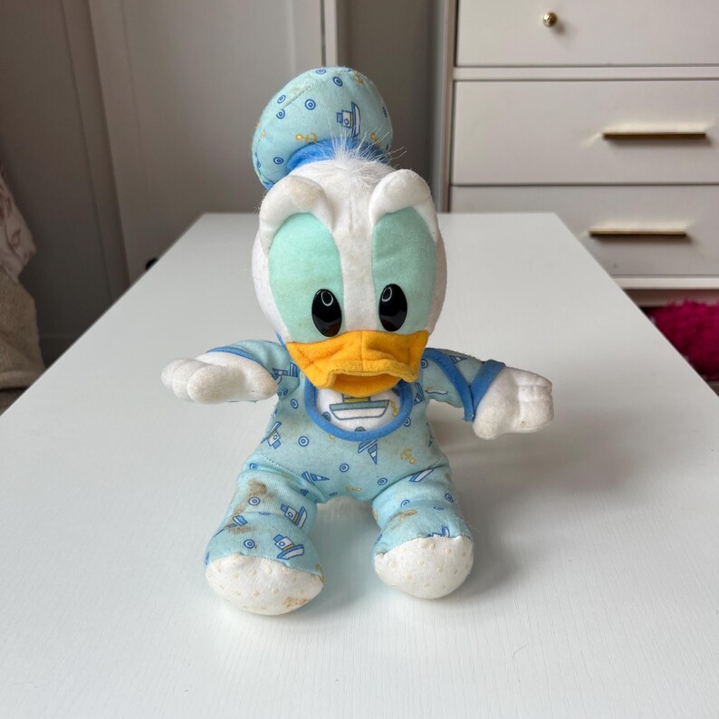 May include: A plush Donald Duck doll in a baby outfit. The doll has a white head with a blue cap, a yellow beak, and black eyes. It wears a light blue onesie with a boat pattern and a bib. The doll is approximately 8 inches tall.