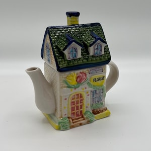 May include: A ceramic teapot shaped like a house, featuring a green roof, blue window frames, and a chimney. The front of the house reads "FLORIST" and has a red door and floral decorations. The teapot is cream-colored.