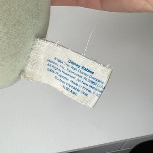 May include: A close-up of a tag from a Disney Babies plush toy. The tag is white with black text, including the Disney Babies logo and manufacturing information. The tag reads "Surface Washable Only."