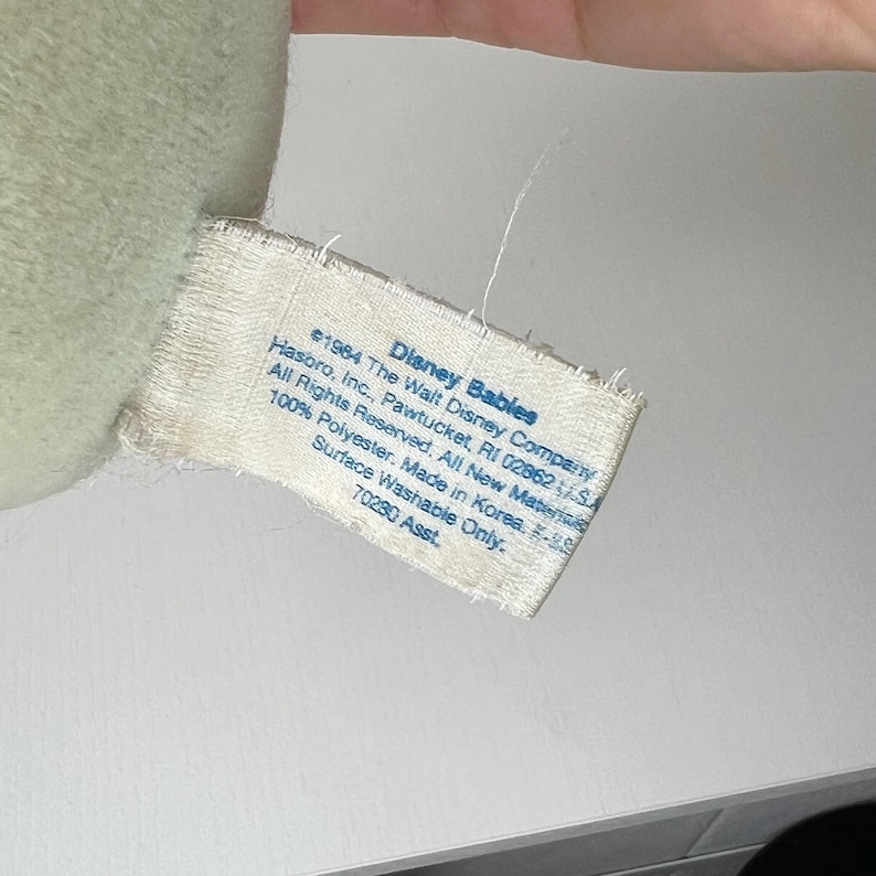 May include: A close-up of a tag from a Disney Babies plush toy. The tag is white with black text, including the Disney Babies logo and manufacturing information. The tag reads "Surface Washable Only."
