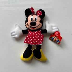 May include: A Minnie Mouse soft toy wearing a red dress with white polka dots and a matching bow. The doll has black ears, arms, and legs, with white gloves and yellow shoes. A red tag with the text "Mickey's Stuff for Kids" is attached.